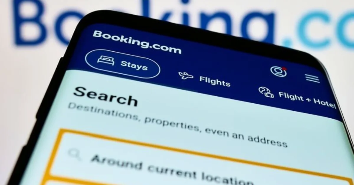 Um close em um smartphone exibindo a tela principal do aplicativo móvel Booking.com com a guia 'Hospedagens' selecionada e a barra de pesquisa visível.