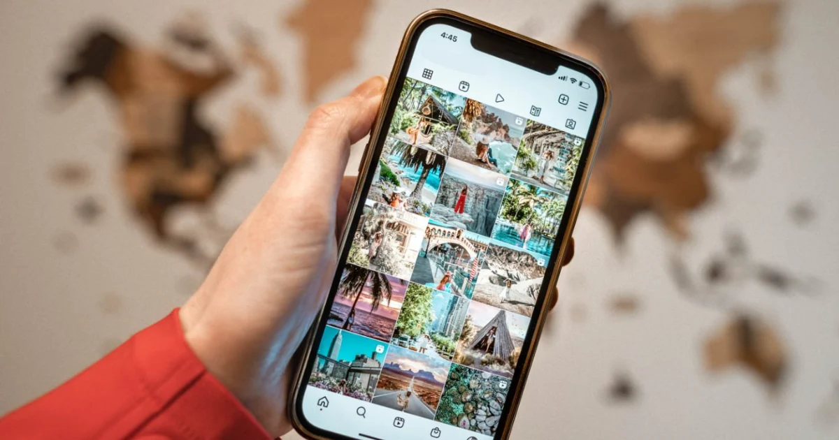Mão segurando um smartphone que exibe um feed do Instagram repleto de fotos de viagens, pousadas e paisagens turísticas, com um mapa-múndi de madeira desfocado ao fundo.