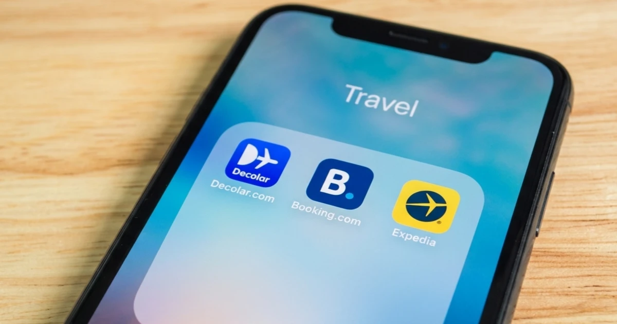 Mãos seguram um smartphone de onde flutuam três telas holográficas com interfaces de aplicativos de viagem: Booking.com, Decolar e Expedia.