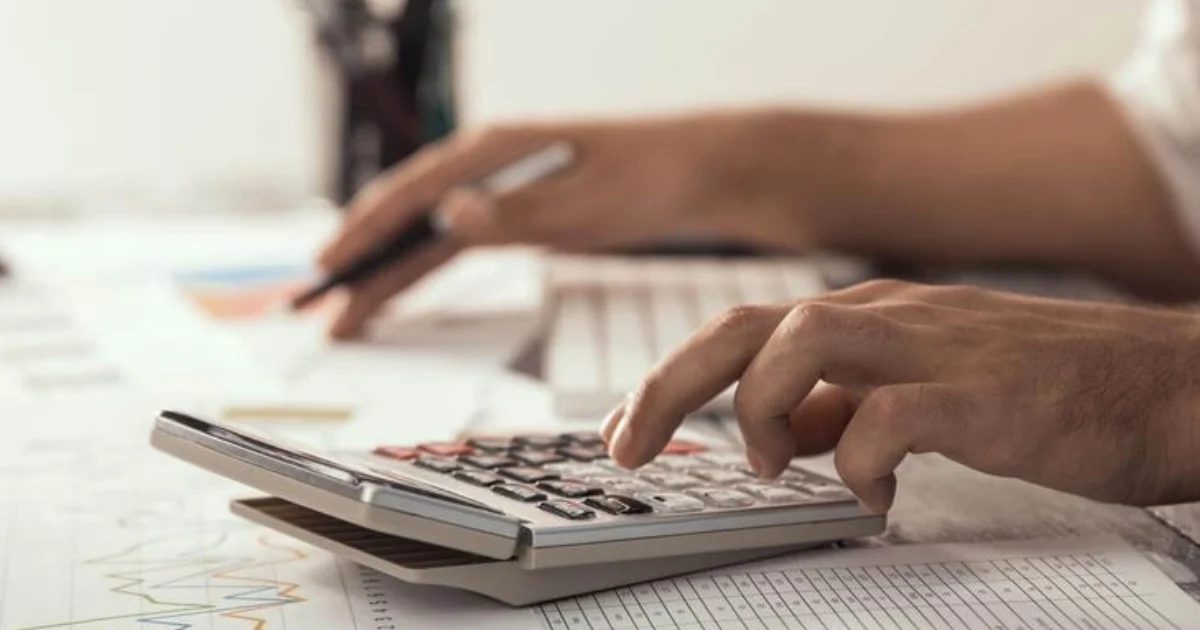 Mão em primeiro plano utilizando uma calculadora sobre planilhas financeiras impressas, com outra pessoa ao fundo segurando uma caneta.