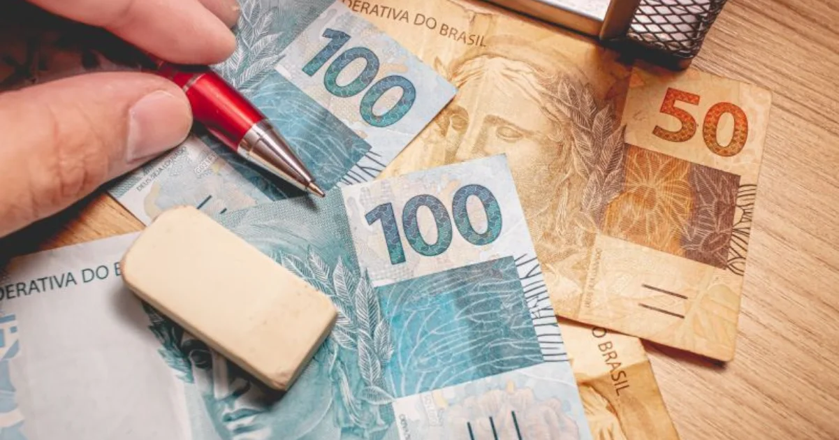 Mão com caneta vermelha sobre notas de cem e cinquenta reais e uma borracha, simbolizando a análise e o cálculo financeiro real de uma hospedagem.