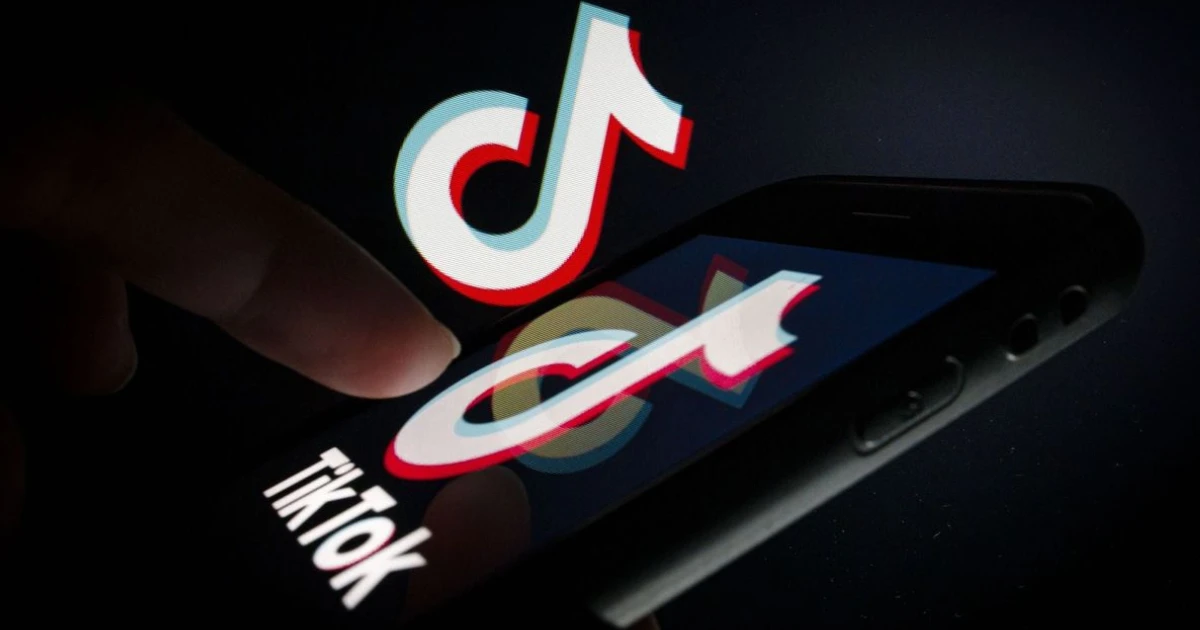 Dedo de um usuário tocando a tela de um smartphone que exibe o logotipo do TikTok em um ambiente escuro.